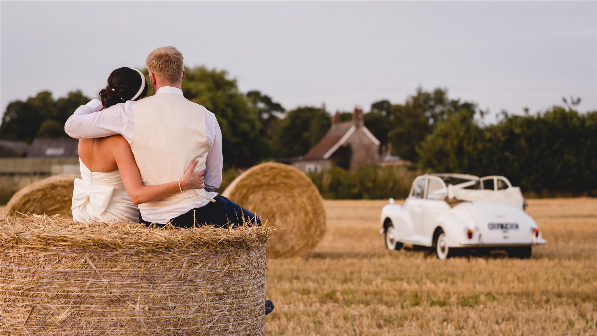 Bride and groom sitting on a hay bale beside a Morris Minor wedding car
