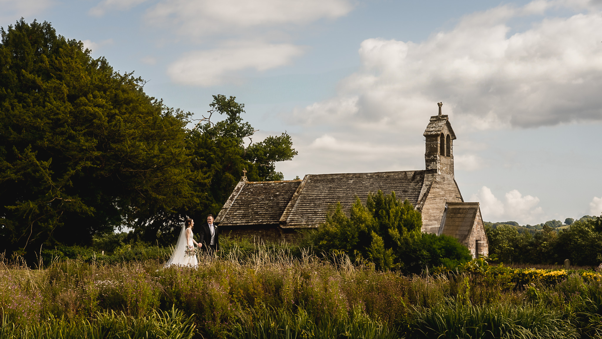 Couple walking through a meadow towards a small stone church in Monmouthshire