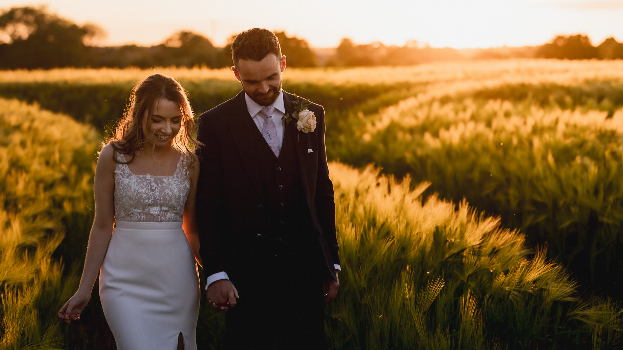 Rachel and Cameron walking through a barley field beside the Great Tythe Barn at golden hour