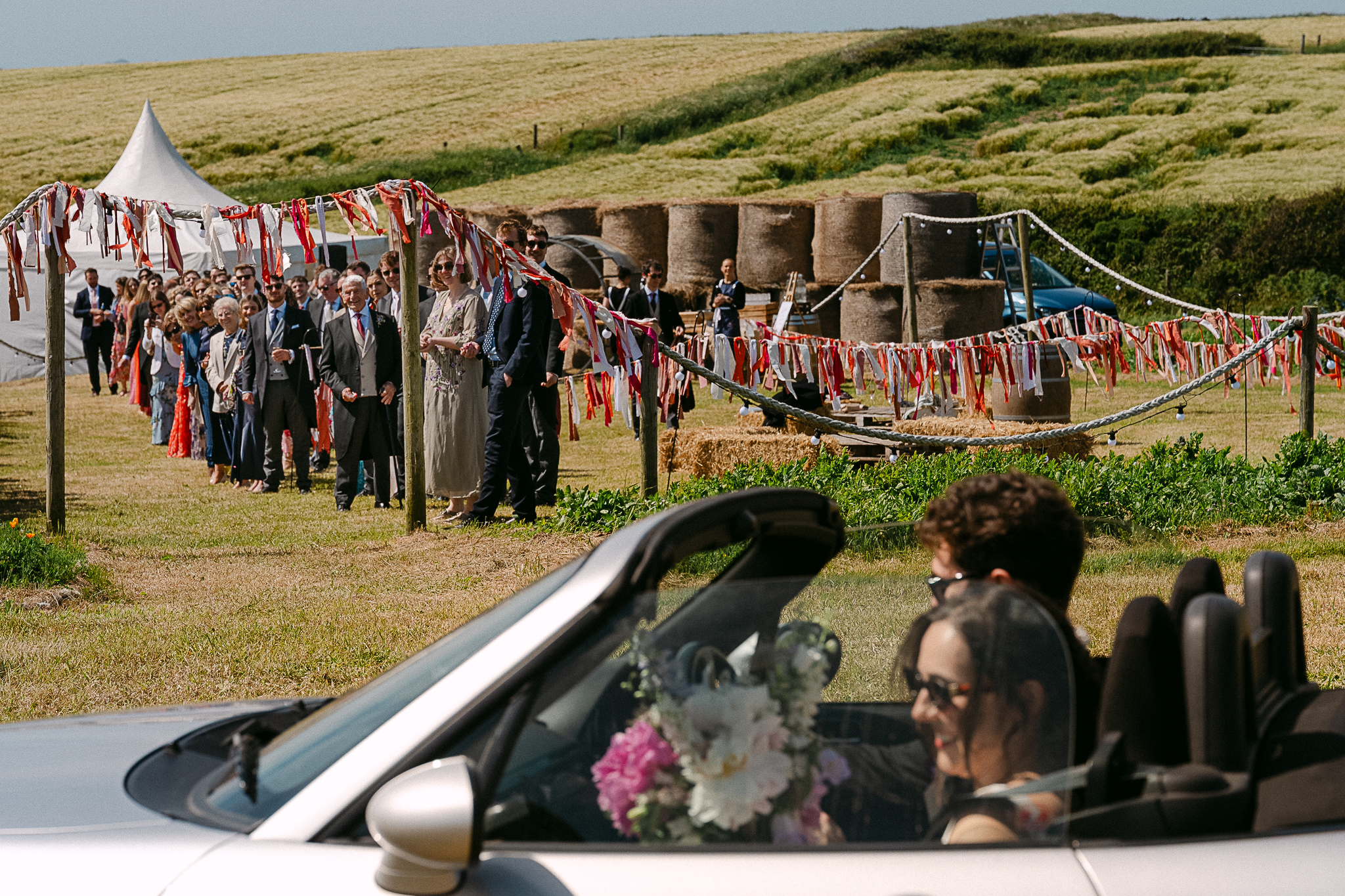 Guests in a vintage convertible arriving at a coastal wedding with bunting and a small tent