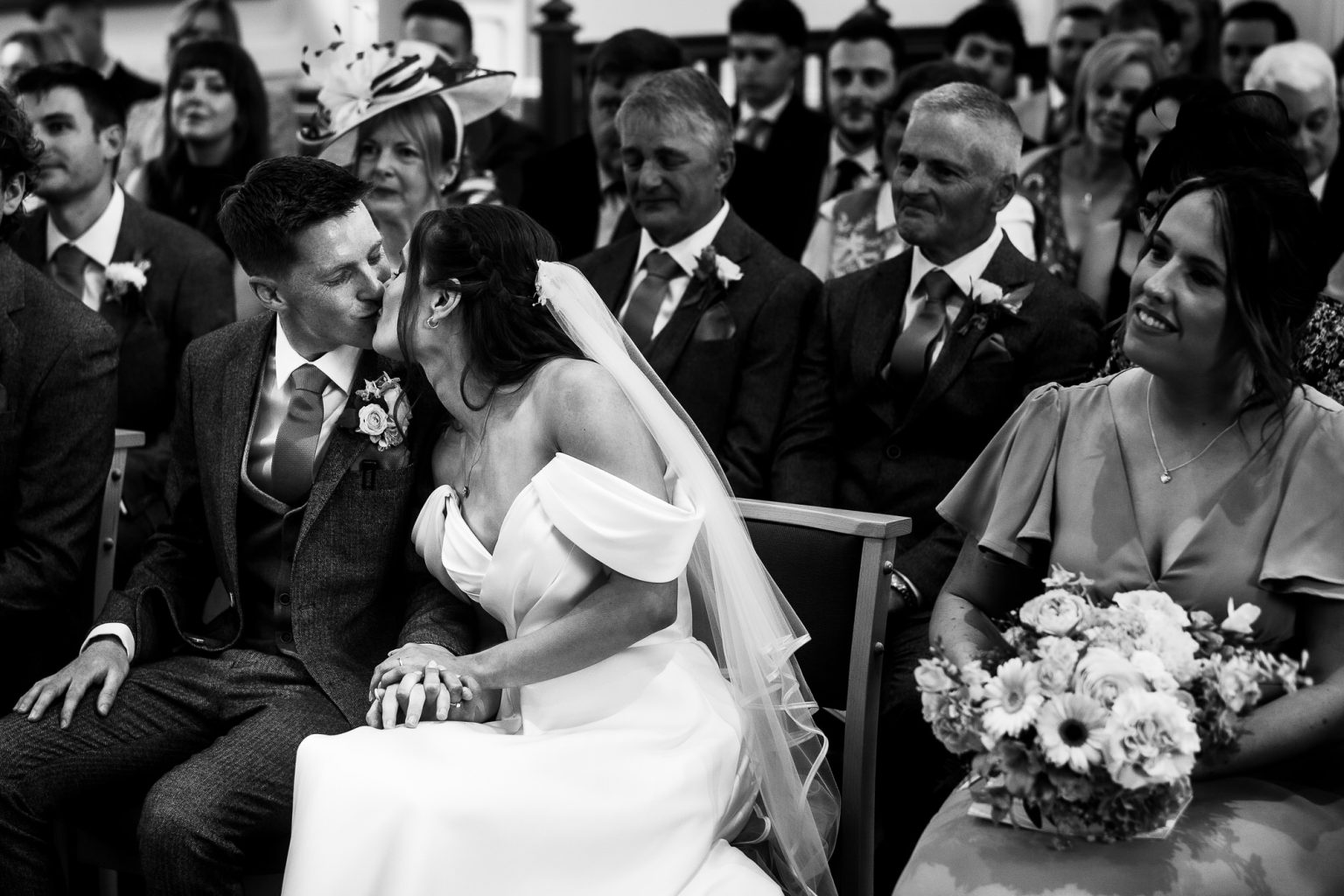 Black and white photo of a couple kissing during their ceremony as family look on