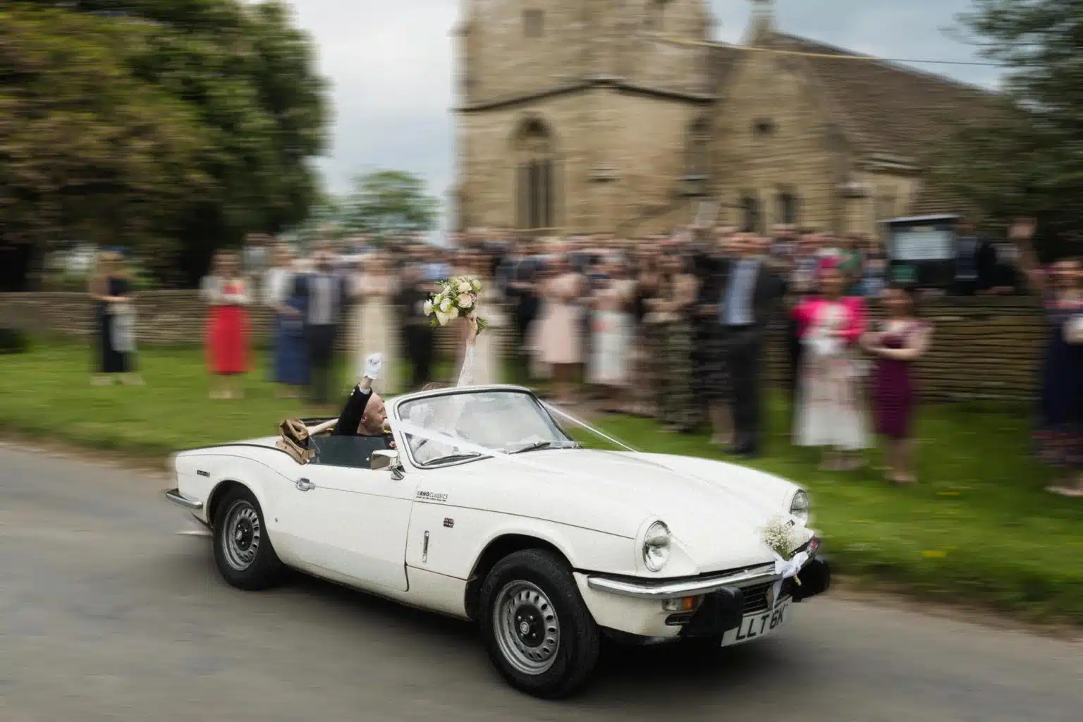 Newlyweds waving from a white vintage convertible as guests cheer outside a village church