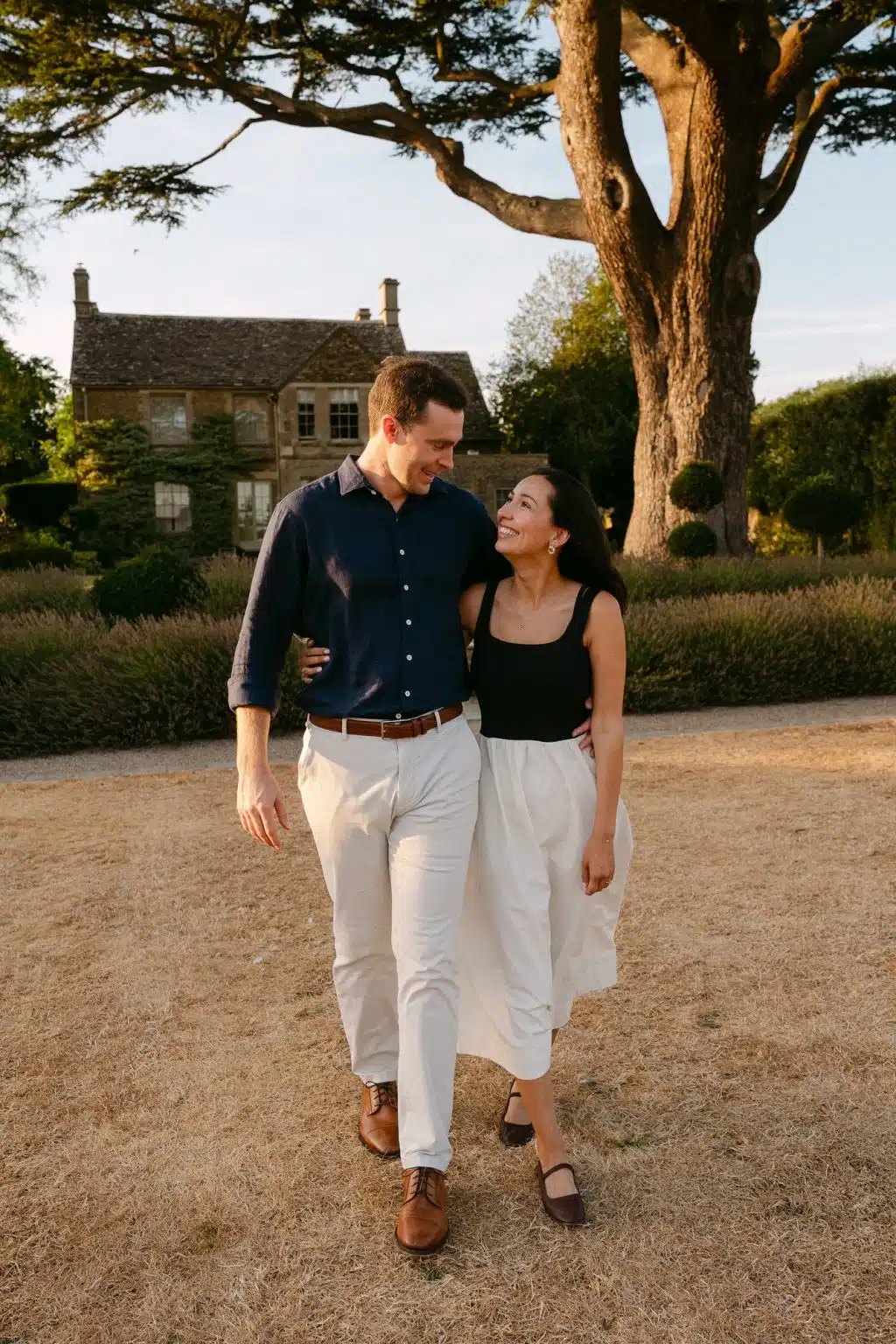 Engaged couple walking arm in arm in front of a Cotswold stone house at golden hour