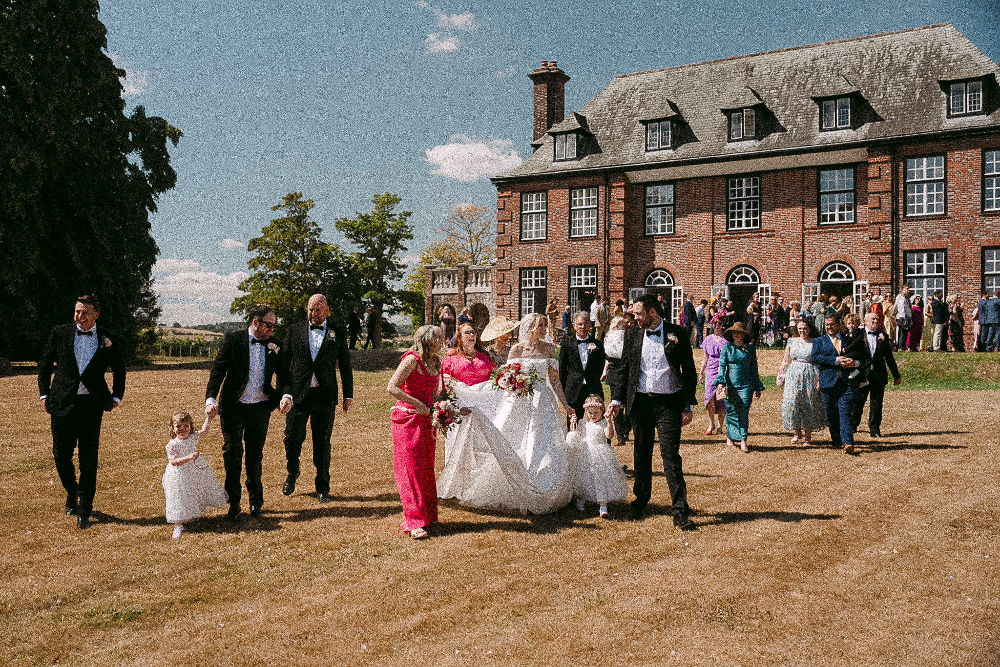 Kate and Dylan walking across the lawn at Sant Ffraed House with their wedding party behind them
