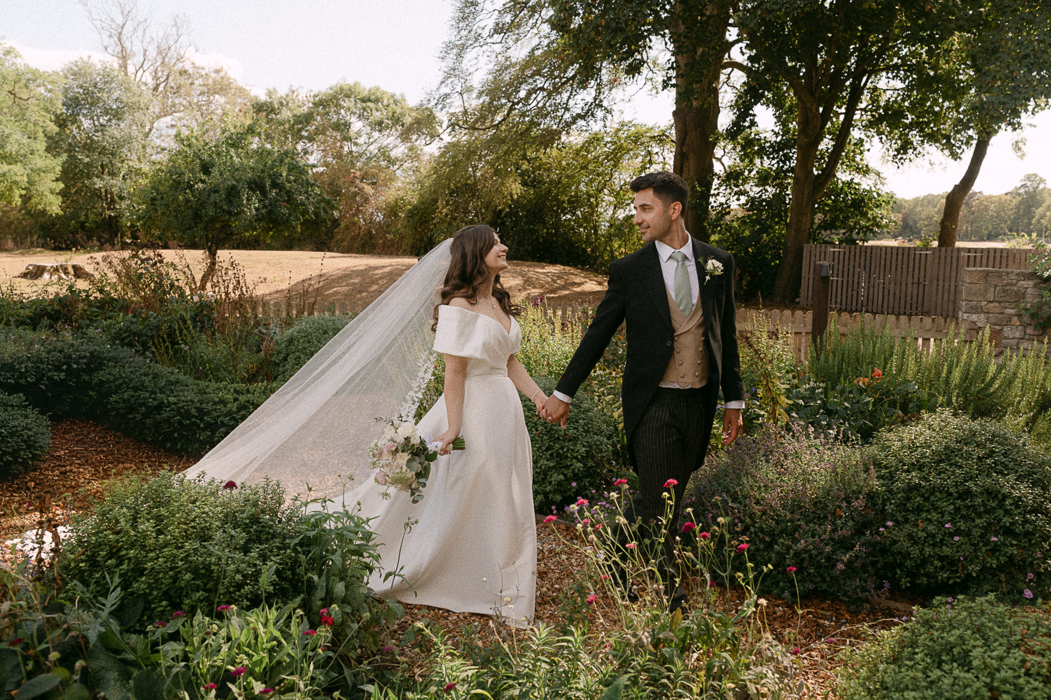 Bride and groom walking through a cottage garden, her veil lifted by the breeze