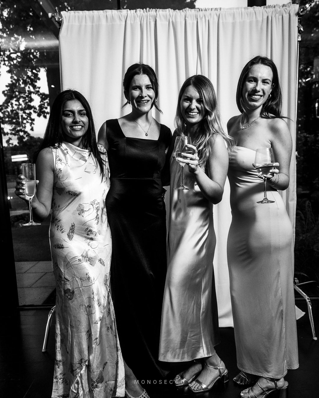 Four friends in long dresses holding drinks in front of the backdrop
