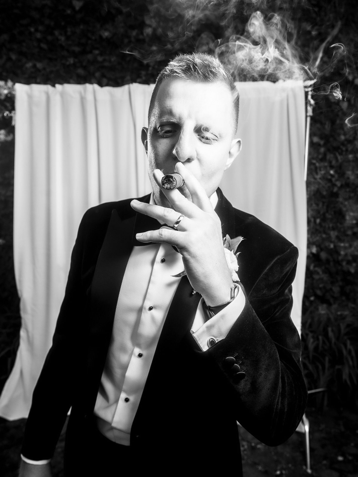 The groom with a cigar in front of a white backdrop at night