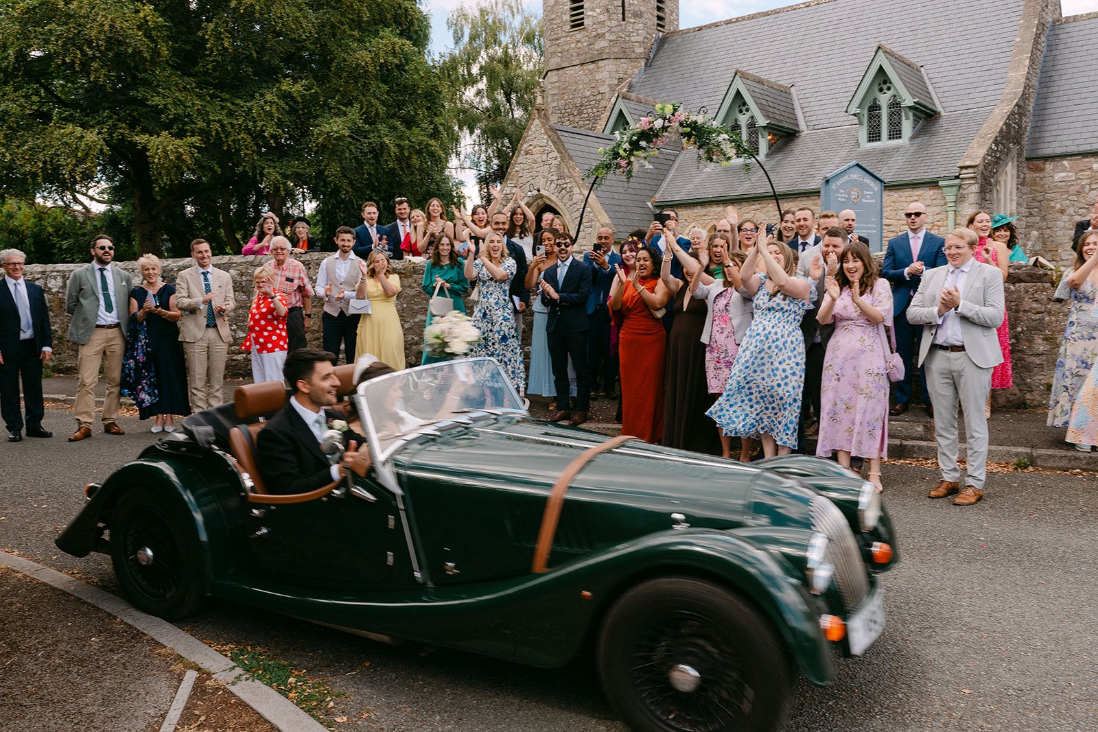 Jess and Tom leaving in an open-top car as guests cheer
